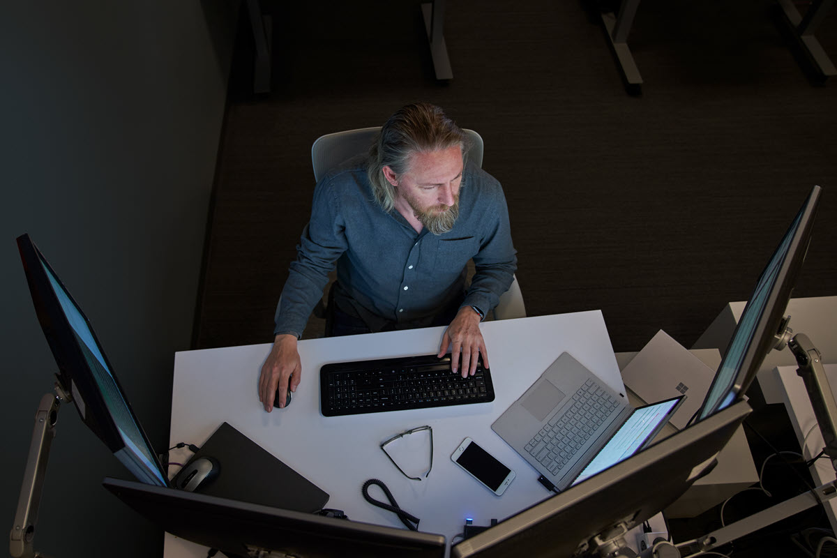 Top-down view of a bearded man in a gray/blue shirt seated at a desk working on a Surface laptop connected to three large monitors