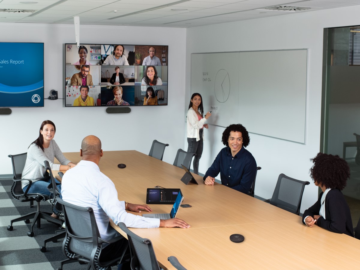 Two males and three females collaborating in a conference room featuring an HP Teams Meeting Rooms touch display. The meeting has nine remote participants shown on a wall mounted display joined via Teams Meetings. Two subjects are using Surface devices