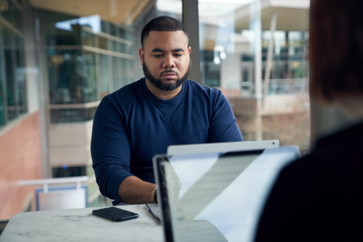 Man sitting at a community table in common area typing on a Surface laptop with a phone at his side. Blurred laptop in foreground