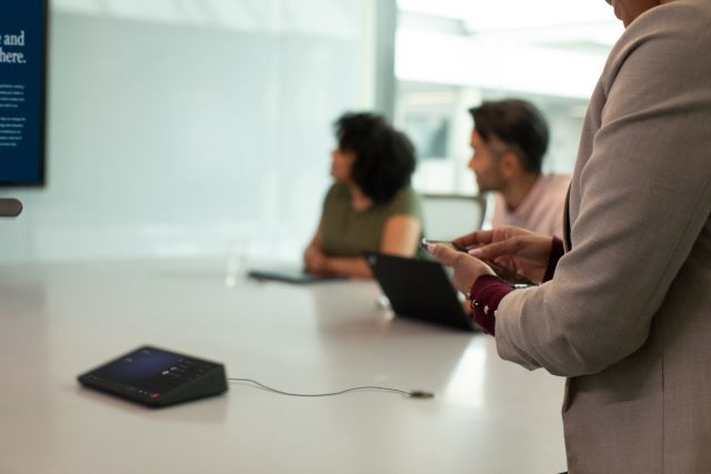 Two women and one man join an in-person meeting in a medium sized conference room with a companion device. One woman is casting from her mobile device while standing at the back of the room