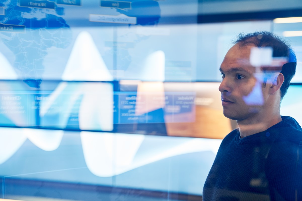 Man in sweater inside a secure room who is looking at data and a geographic area displayed on a large monitor which is behind glass walls with reflections