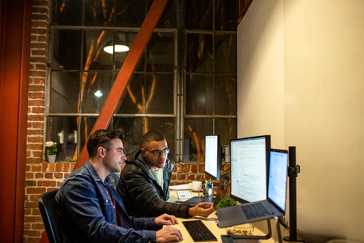 Photo of two male developers working and collaborating in an casual enterprise office. They are using Visual Studio with a multi-monitor set up