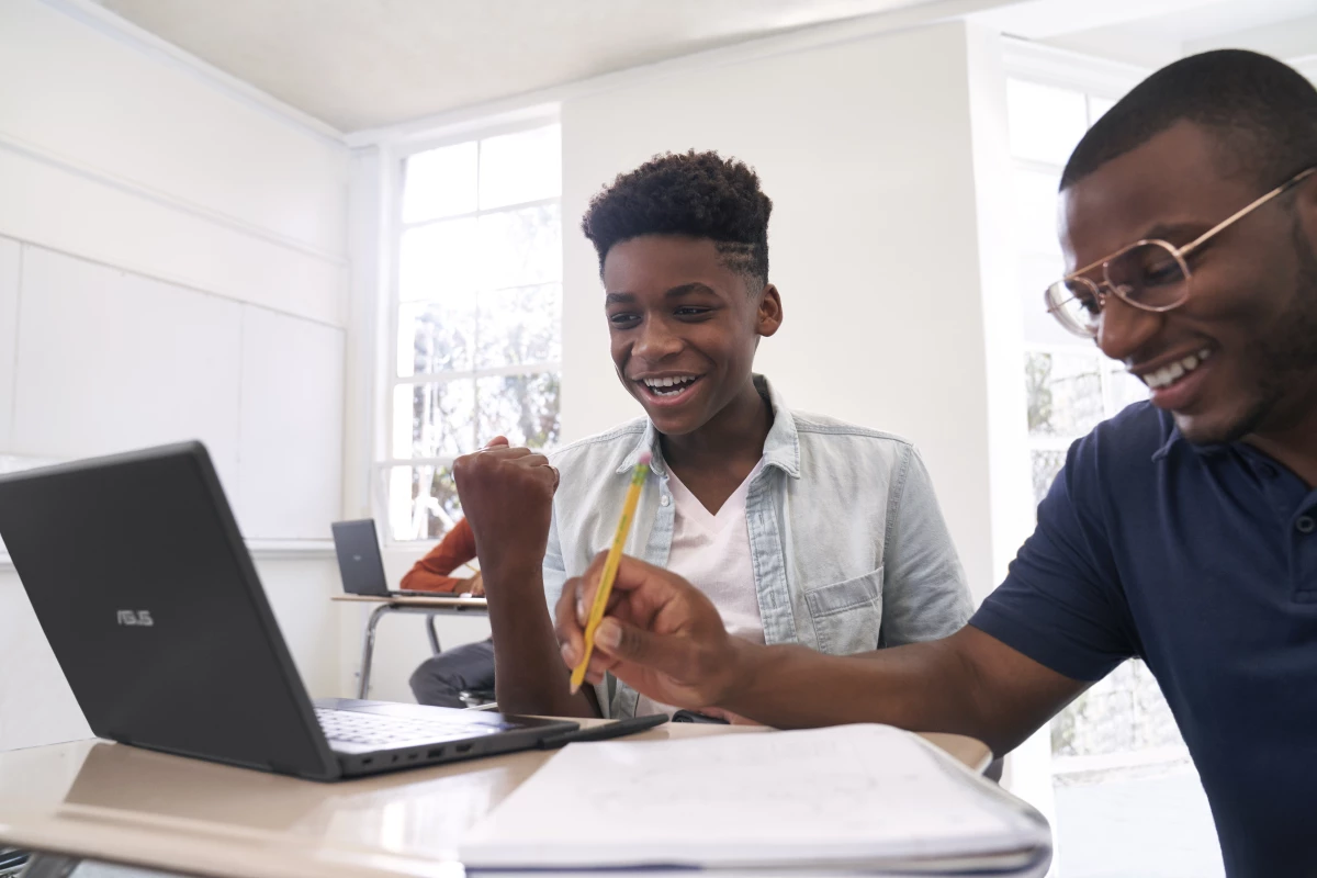 Excited male K-12 student sitting at a classroom desk using an ASUS BR1100 CKA device. Smiling male educator kneeling beside him