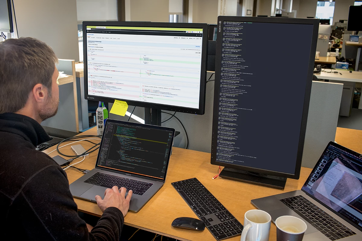 Photo of Male developer is coding at his workspace in an enterprise office using Visual Studio on a MacBook Pro multi-monitor set up