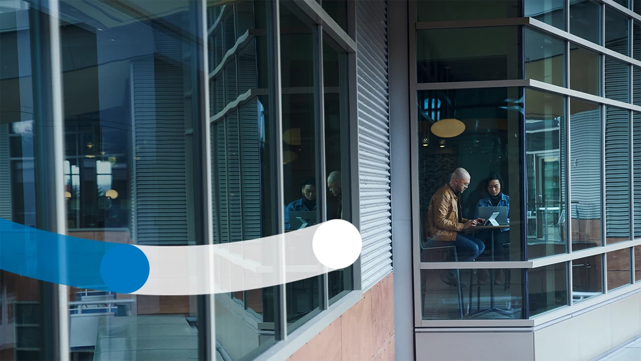 Looking through the windows from the outside of a multi-level building at a man and woman working on their laptops together at a high table
