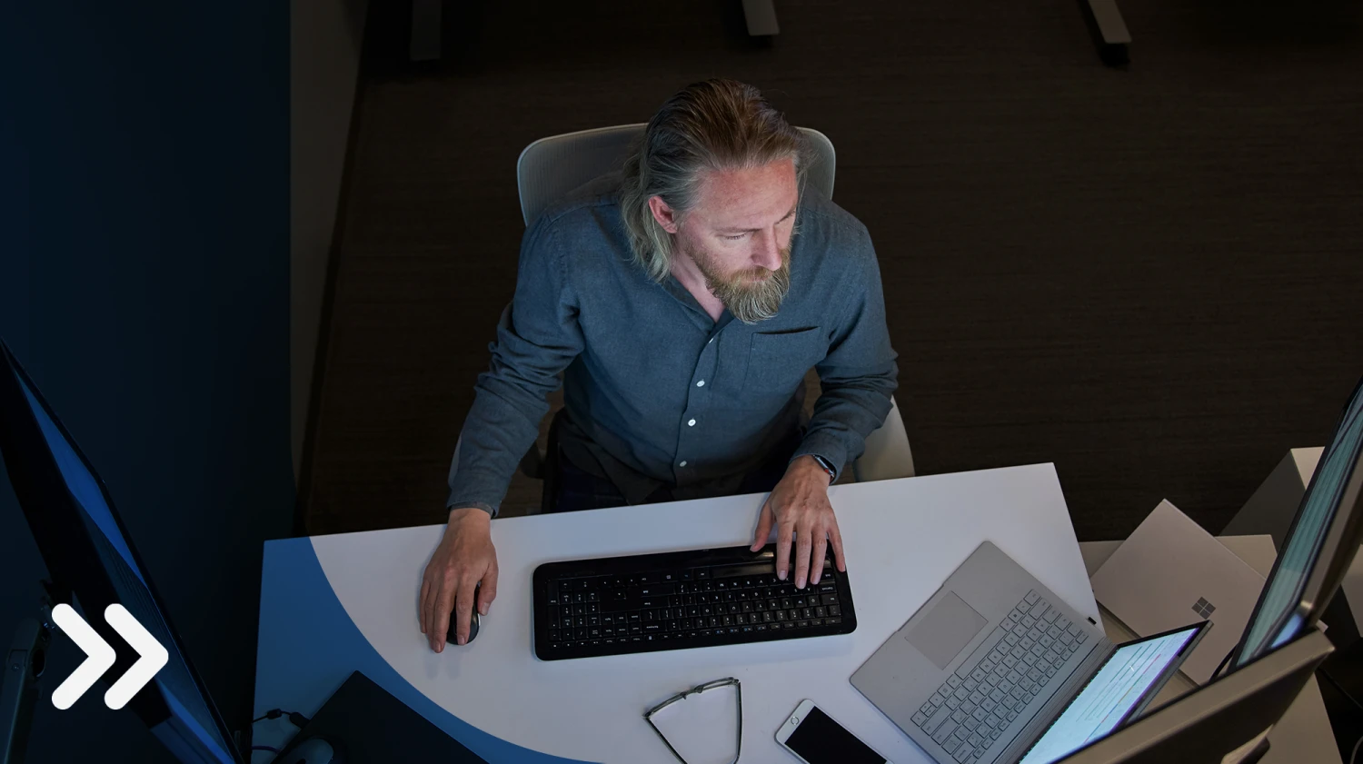 Top-down view of a bearded man in a gray/blue shirt seated at a desk working on a Surface laptop connected to three large monitors. Desktop has phone, glasses, mouse and other laptops on it