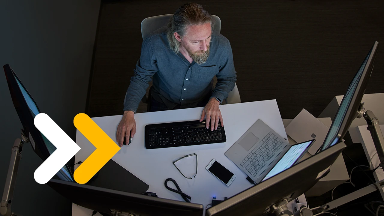 Top-down view of a bearded man in a gray/blue shirt seated at a desk working on a Surface laptop connected to three large monitors